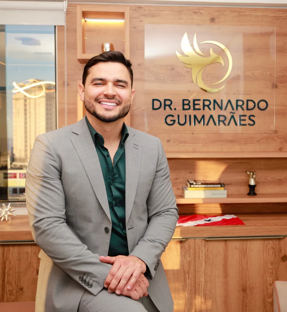 Dr. Bernardo Guimarães sorrindo, de terno cinza, em frente ao painel com o logo do Instituto